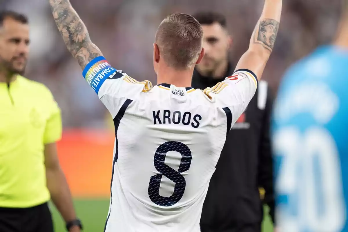 Un jugador de fútbol con el número 8 y el nombre "Kroos" en su camiseta levanta los brazos en el campo, mientras un árbitro y otros jugadores están cerca.