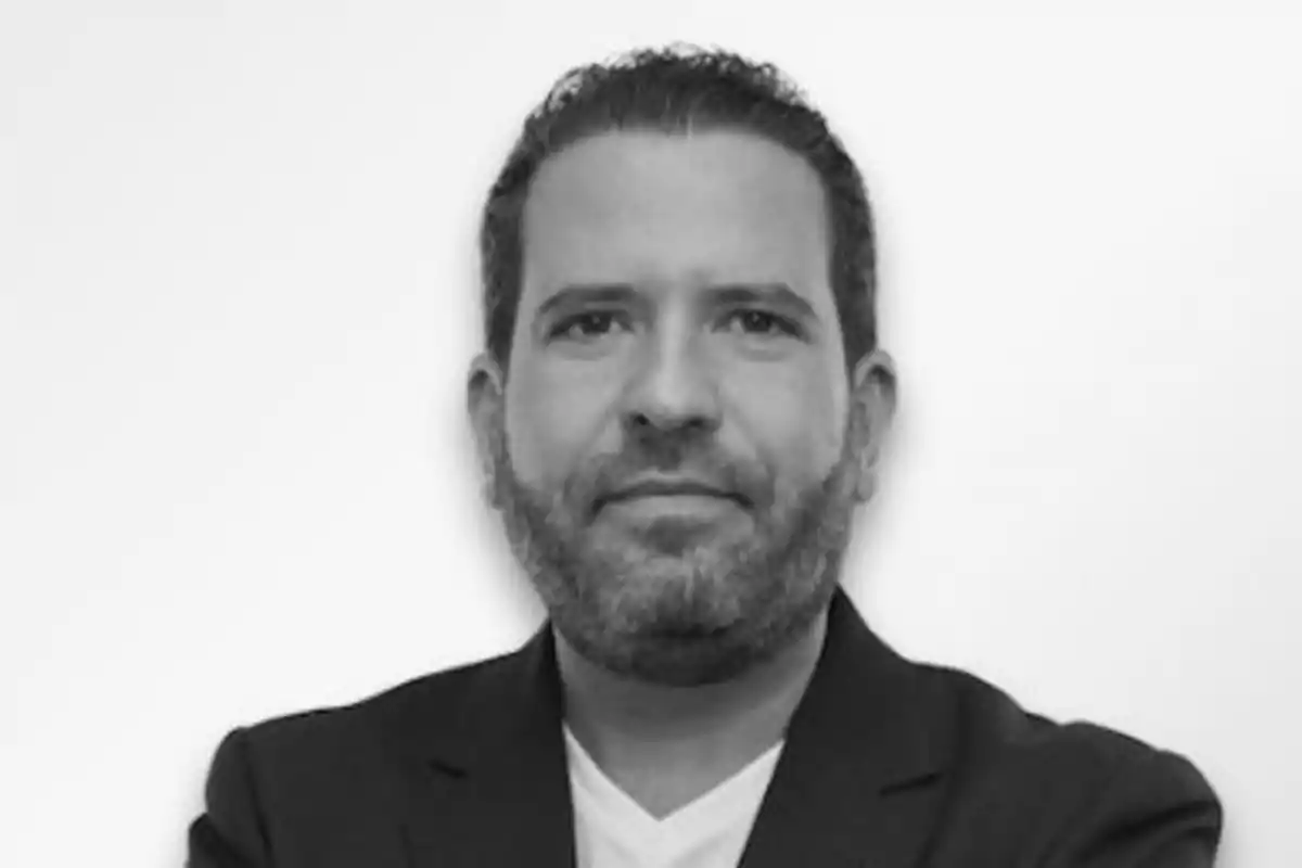 Hombre de cabello corto y barba, vestido con saco oscuro y camiseta clara, mirando al frente con expresión seria sobre fondo blanco Hombre de cabello corto y barba, vestido con saco oscuro y camiseta clara, mirando al frente con expresión seria sobre fondo blanco