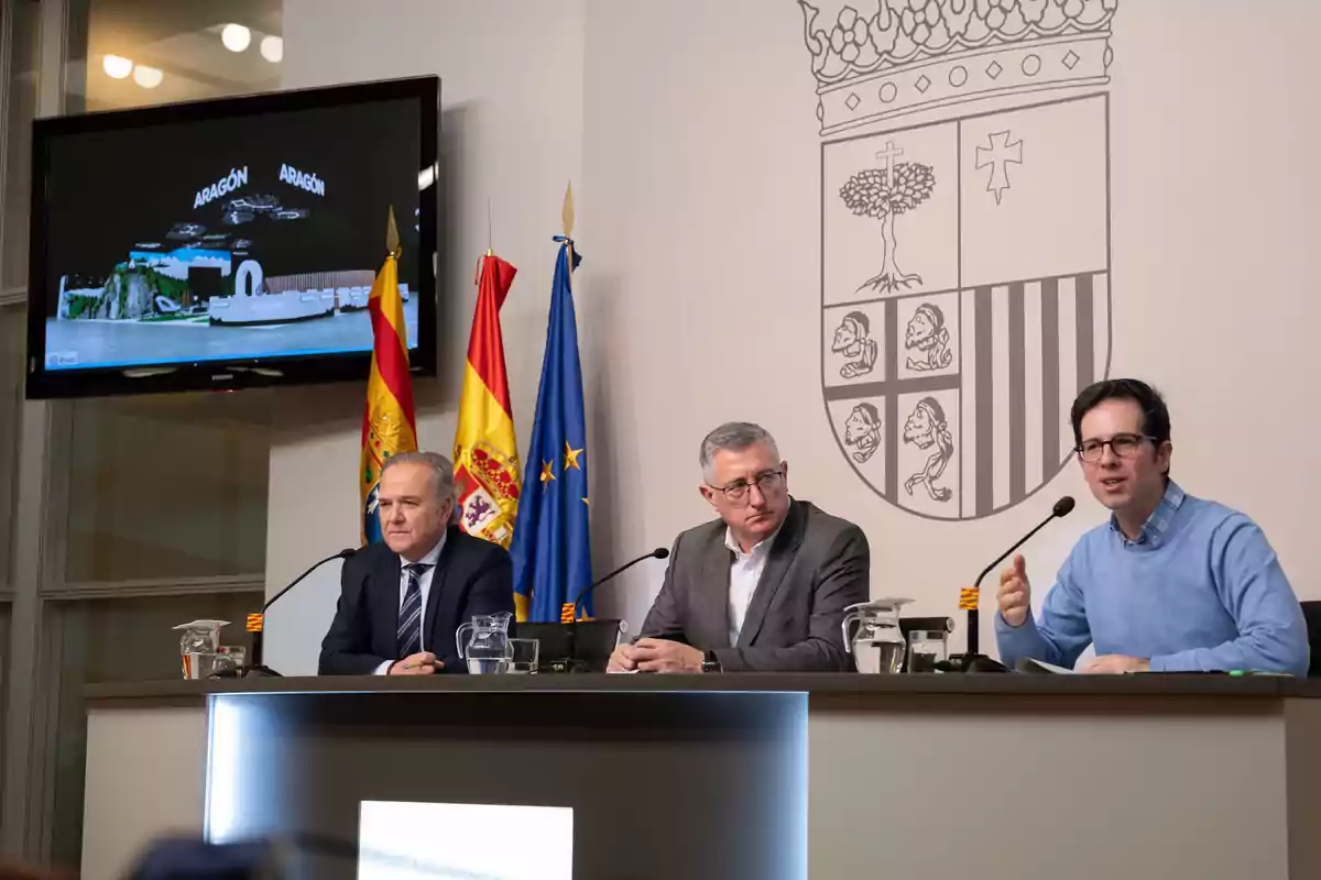 Tres hombres sentados en una mesa durante una conferencia de prensa con banderas de España y Aragón detrás y un escudo en la pared.