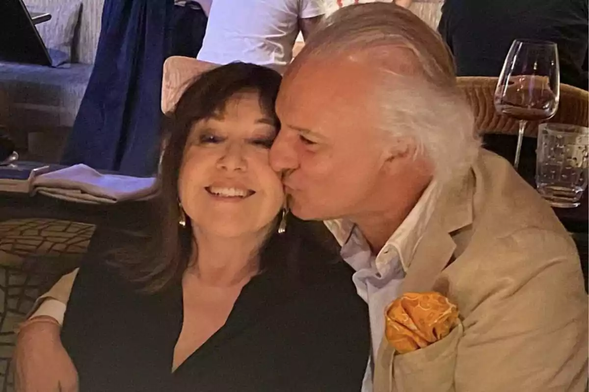 A couple smiles as the man kisses the woman on the cheek in a restaurant setting.