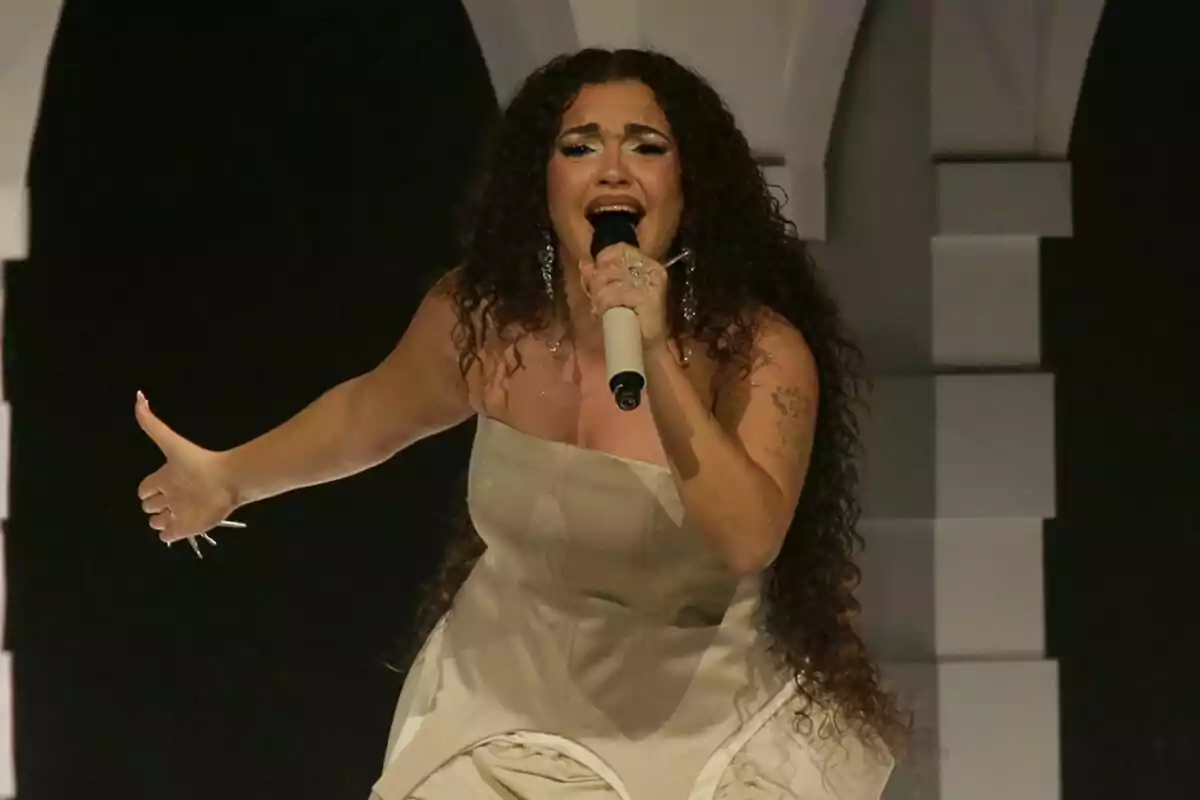 La Chispa con cabello rizado y vestido claro sosteniendo un micrófono mientras canta en un escenario en el Benidorm Fest. La Chispa con cabello rizado y vestido claro sosteniendo un micrófono mientras canta en un escenario en el Benidorm Fest.