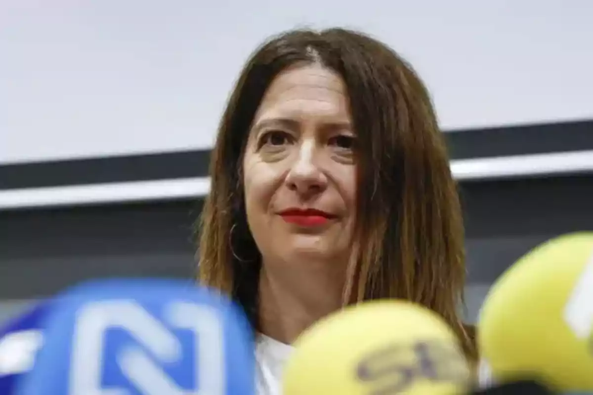 Mujer de cabello castaño largo y labios rojos hablando frente a varios micrófonos de colores en una conferencia de prensa Mujer de cabello castaño largo y labios rojos hablando frente a varios micrófonos de colores en una conferencia de prensa