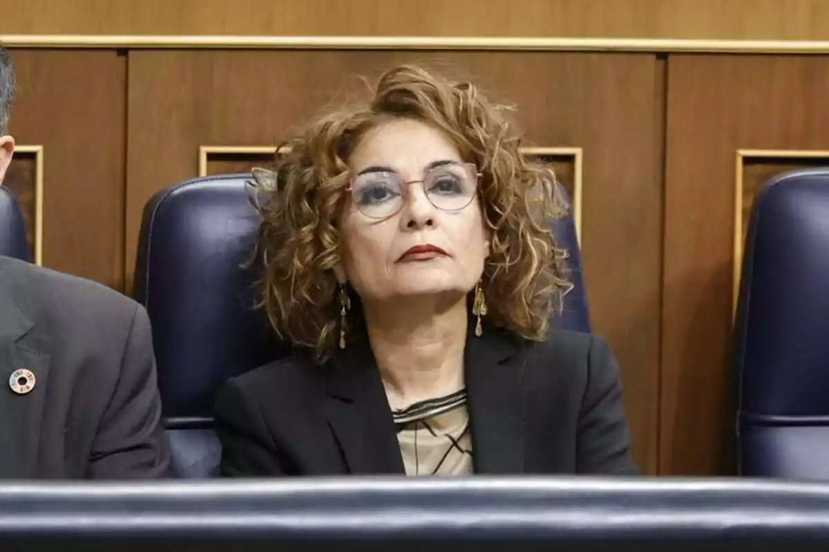 Una mujer con gafas y cabello rizado está sentada en un asiento azul en un entorno formal.