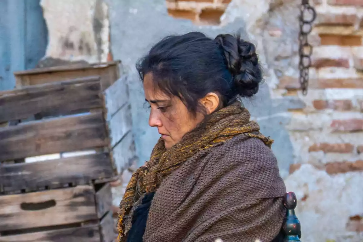 Alicia con cabello recogido y bufanda marrón, de perfil, frente a un fondo de cajas de madera y pared de ladrillo. Captura de 'La Promesa' Alicia con cabello recogido y bufanda marrón, de perfil, frente a un fondo de cajas de madera y pared de ladrillo. Captura de 'La Promesa'