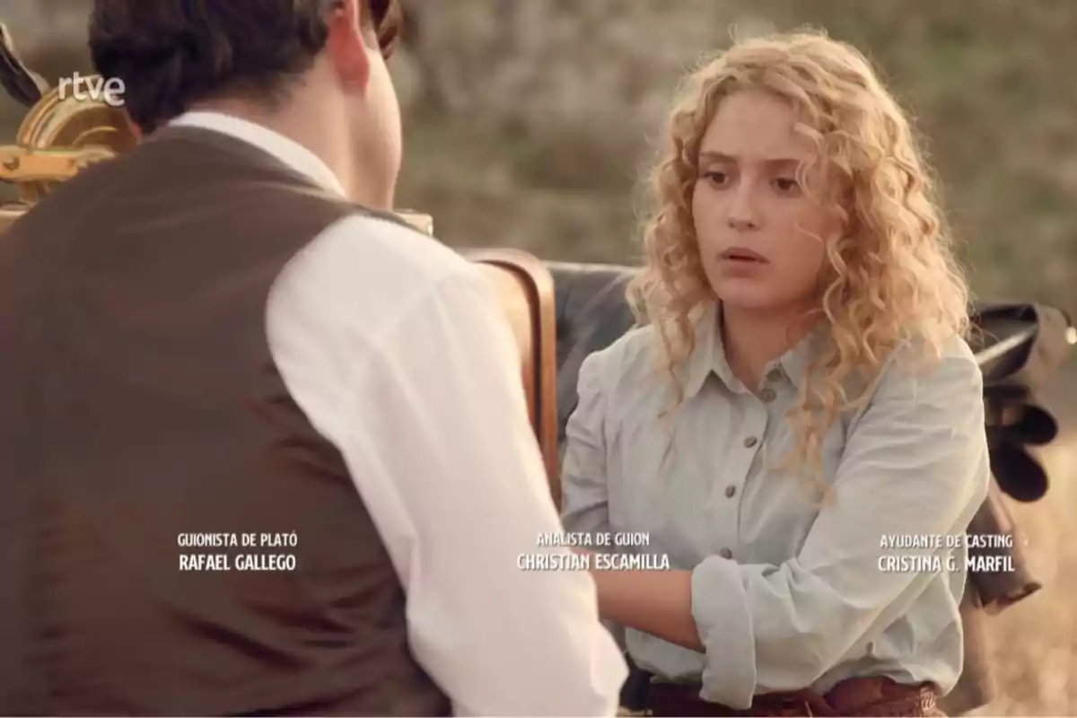 Jana de cabello rizado y expresión seria conversa con Manuel de espaldas en un entorno al aire libre, con créditos de producción visibles en la imagen. Captura de 'La Promesa'