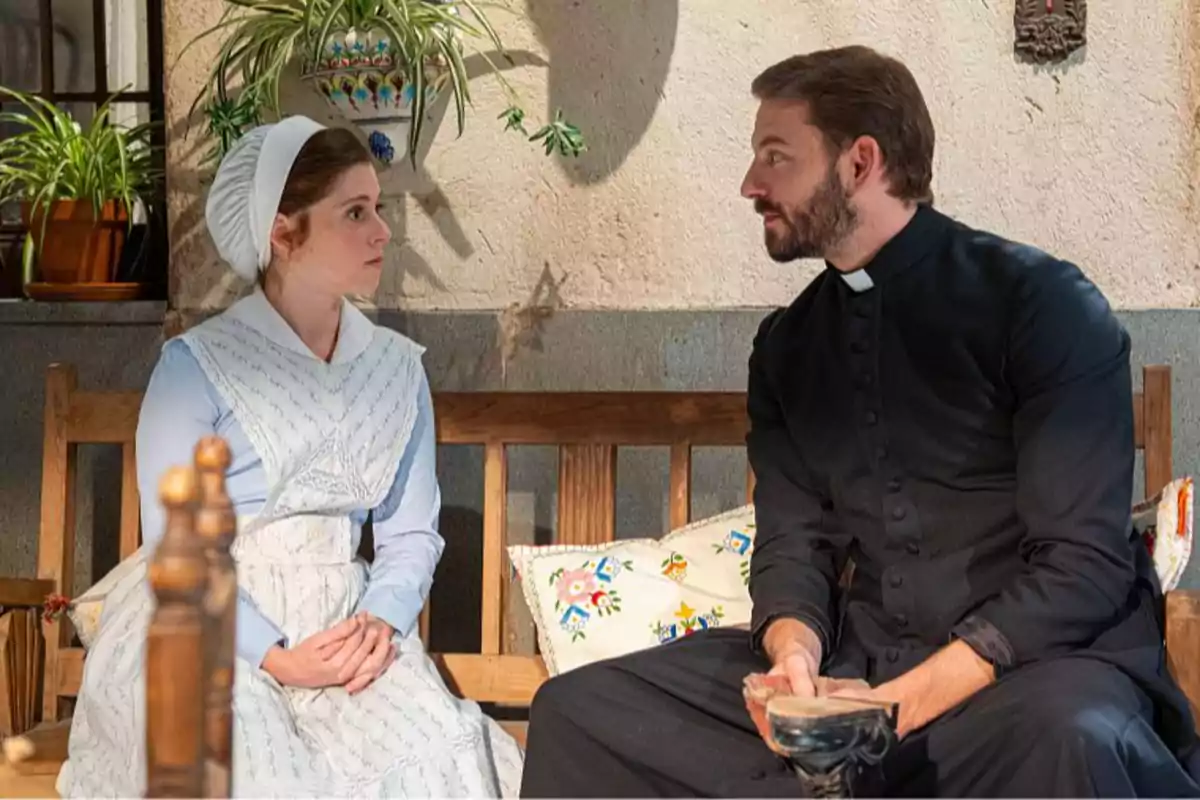 María Fernández vestida de época con cofia blanca y el padre Fernández con sotana negra están sentados en un banco de madera, mirándose y conversando en un ambiente decorado con plantas y cojines coloridos. Captura de 'La Promesa' María Fernández vestida de época con cofia blanca y el padre Fernández con sotana negra están sentados en un banco de madera, mirándose y conversando en un ambiente decorado con plantas y cojines coloridos. Captura de 'La Promesa'