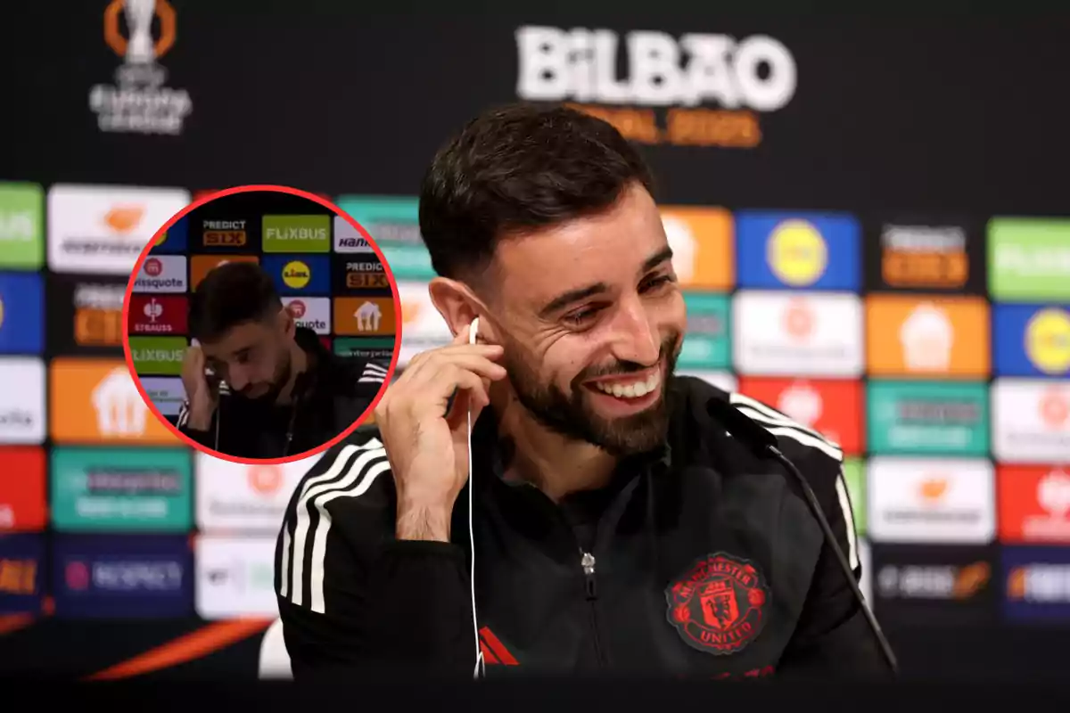 Un jugador de fútbol sonríe durante una conferencia de prensa con un fondo de logotipos de patrocinadores y un micrófono frente a él.