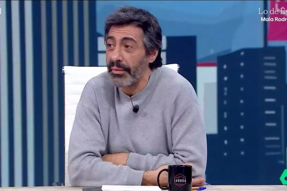 Un hombre con barba y cabello oscuro está sentado en un estudio de televisión, lleva un suéter gris y tiene un micrófono de solapa, frente a él hay una taza negra sobre la mesa. Un hombre con barba y cabello oscuro está sentado en un estudio de televisión, lleva un suéter gris y tiene un micrófono de solapa, frente a él hay una taza negra sobre la mesa.