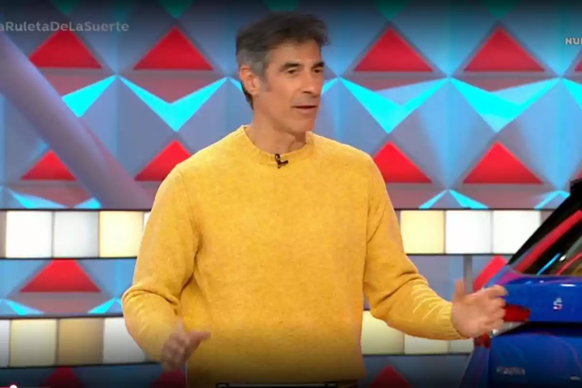 Jorge Fernández in a yellow sweater stands on a colorful TV set with geometric patterns in the background. Still from 'La ruleta de la Suerte'