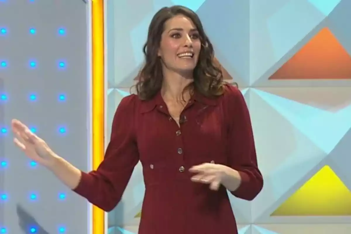 Laura Moure con vestido rojo sonríe mientras gesticula en un set de televisión con fondo geométrico y luces azules. Captura de 'La Ruleta de la Suerte' Laura Moure con vestido rojo sonríe mientras gesticula en un set de televisión con fondo geométrico y luces azules. Captura de 'La Ruleta de la Suerte'