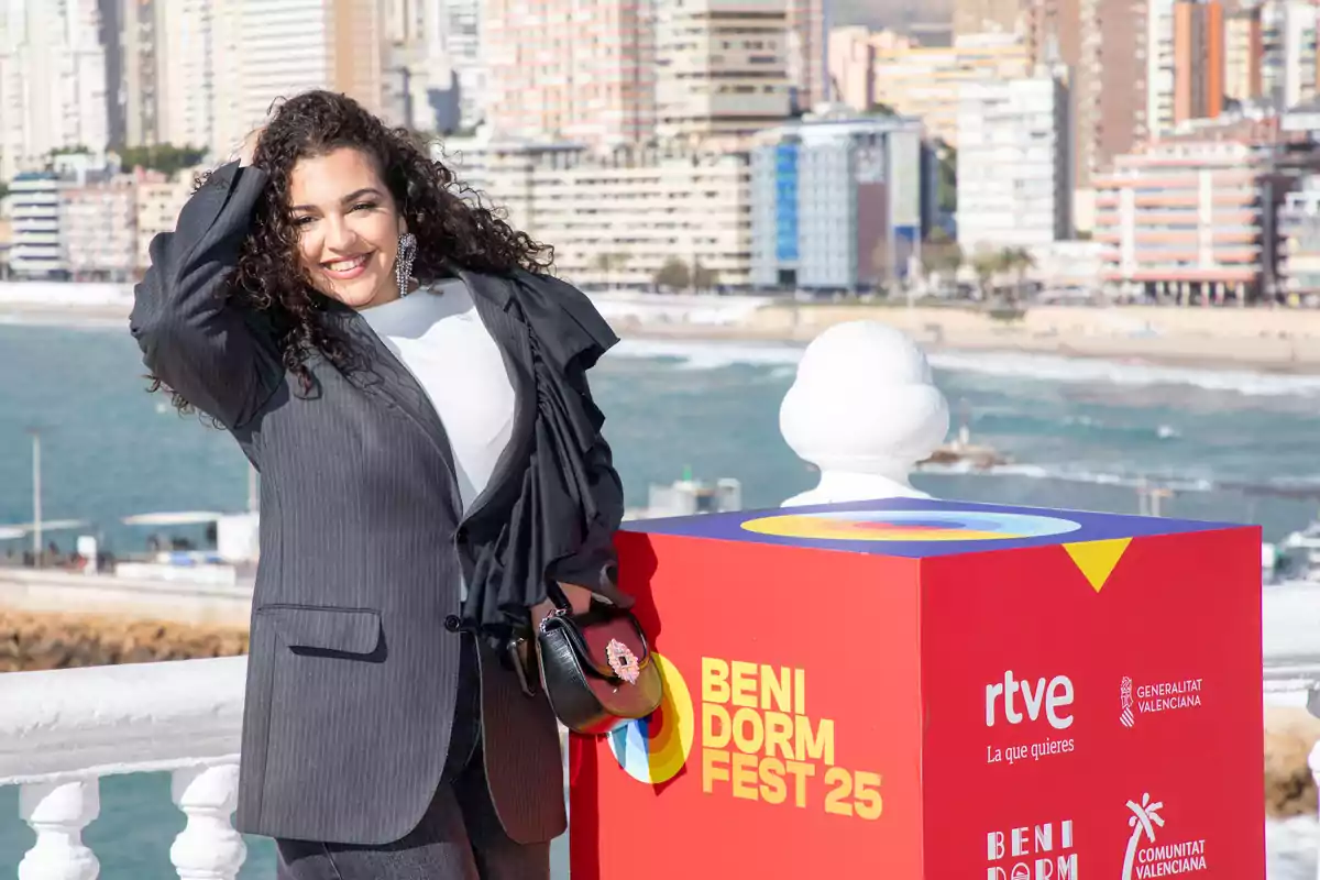 Lachispa sonriente posa junto a un cartel del Benidorm Fest 25 con el mar y edificios de fondo. Lachispa sonriente posa junto a un cartel del Benidorm Fest 25 con el mar y edificios de fondo.