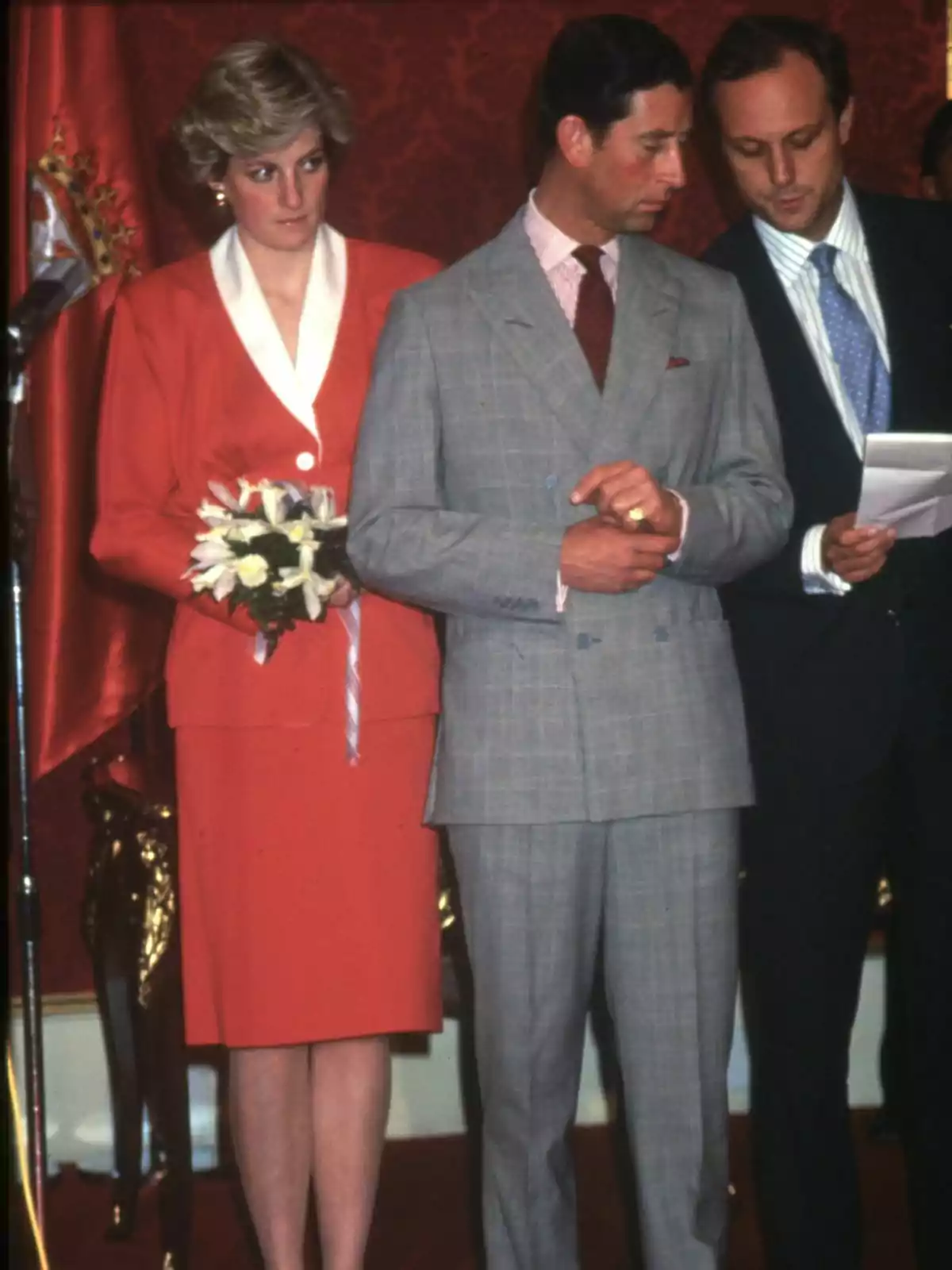 Lady Di con un ramo de flores y Carlos III de traje están de pie en un evento formal. Lady Di con un ramo de flores y Carlos III de traje están de pie en un evento formal.