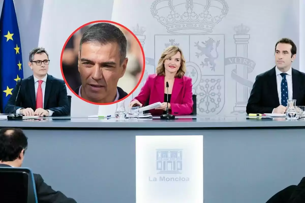 Personas en una conferencia de prensa en La Moncloa con una bandera de la Unión Europea al fondo y un retrato enmarcado de un hombre en el centro.