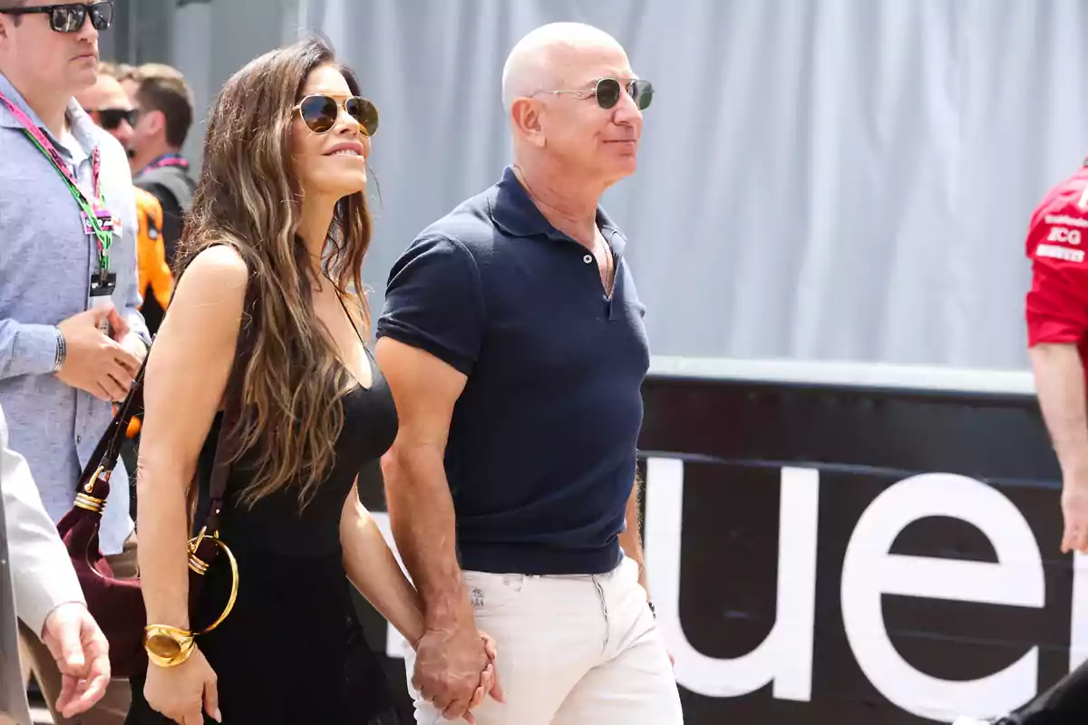 Lauren Sánchez and Jeff Bezos walking hand in hand outdoors, both wearing sunglasses and casual clothes