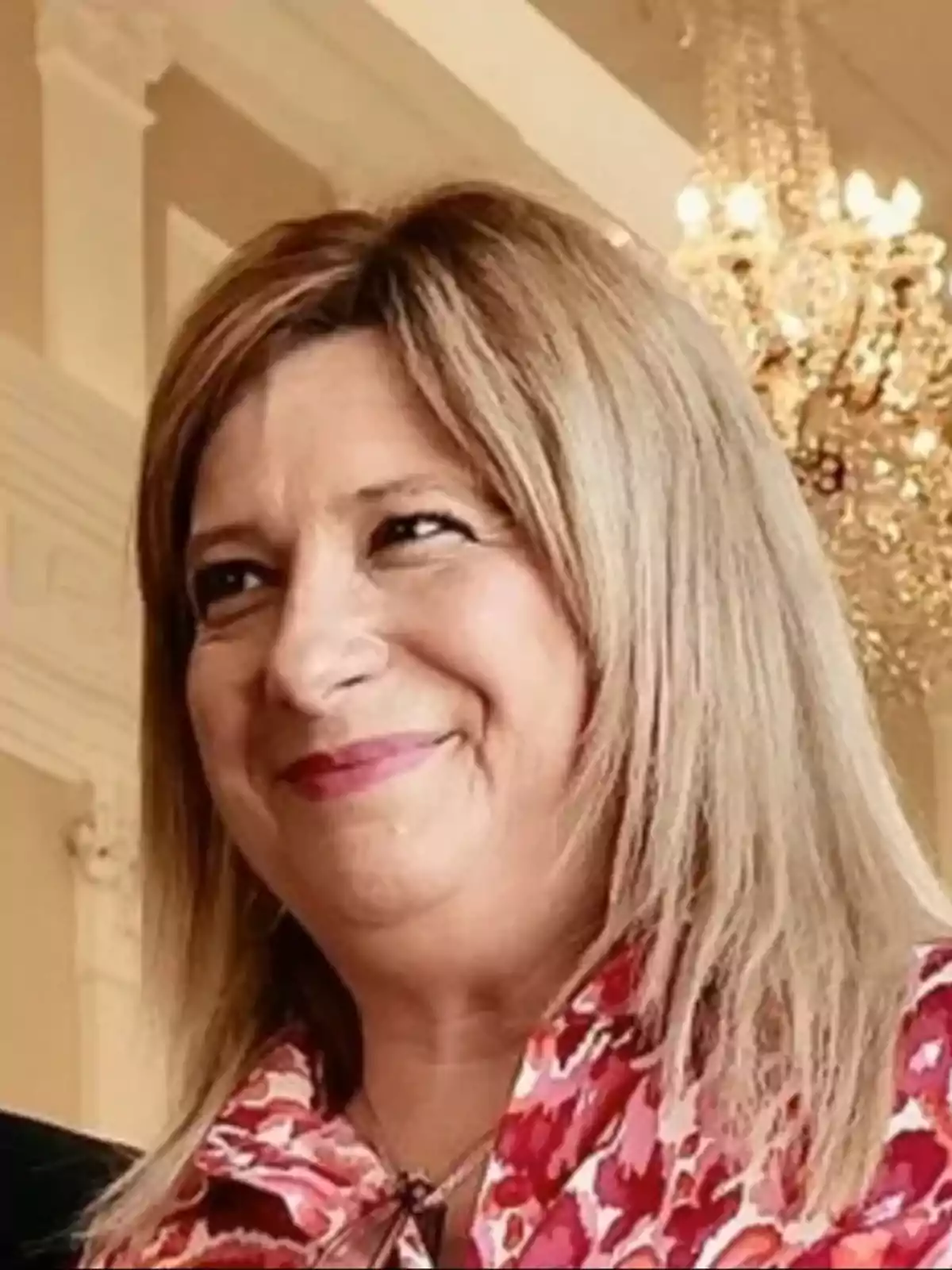 Mujer de cabello rubio sonriente con blusa de estampado rojo en un ambiente elegante con lámpara de araña al fondo Mujer de cabello rubio sonriente con blusa de estampado rojo en un ambiente elegante con lámpara de araña al fondo