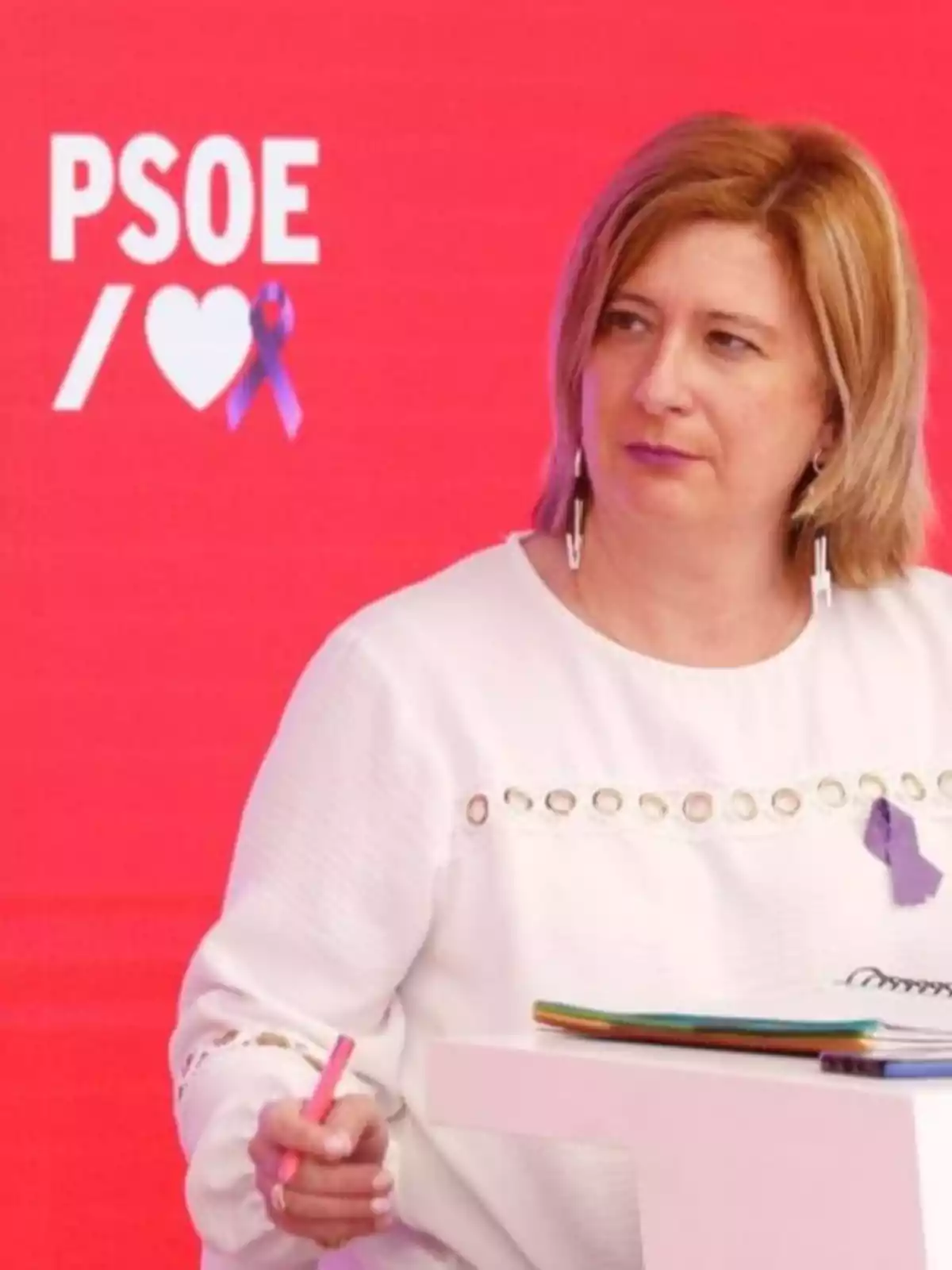 Mujer de cabello rubio y corto vestida con blusa blanca y lazo morado sentada frente a un fondo rojo con el logo de PSOE y un corazón blanco Mujer de cabello rubio y corto vestida con blusa blanca y lazo morado sentada frente a un fondo rojo con el logo de PSOE y un corazón blanco
