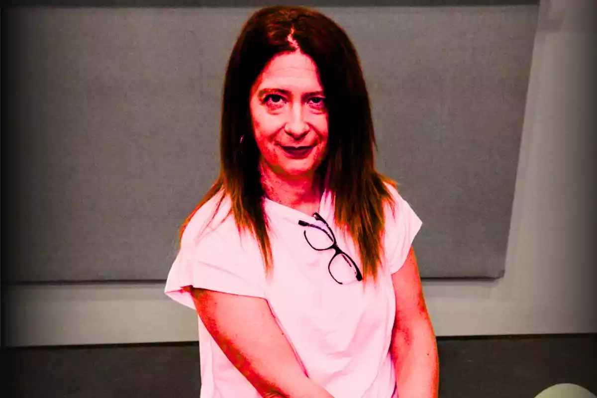 Mujer de cabello largo y lacio con camiseta clara y gafas colgadas en el cuello, sentada frente a un fondo gris Mujer de cabello largo y lacio con camiseta clara y gafas colgadas en el cuello, sentada frente a un fondo gris