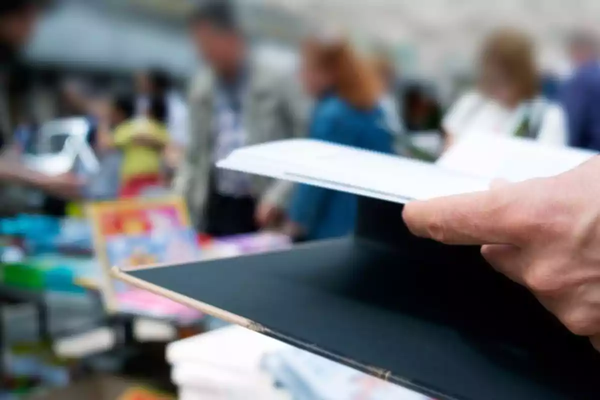 Una persona hojea un libro en un mercado al aire libre con gente desenfocada al fondo.