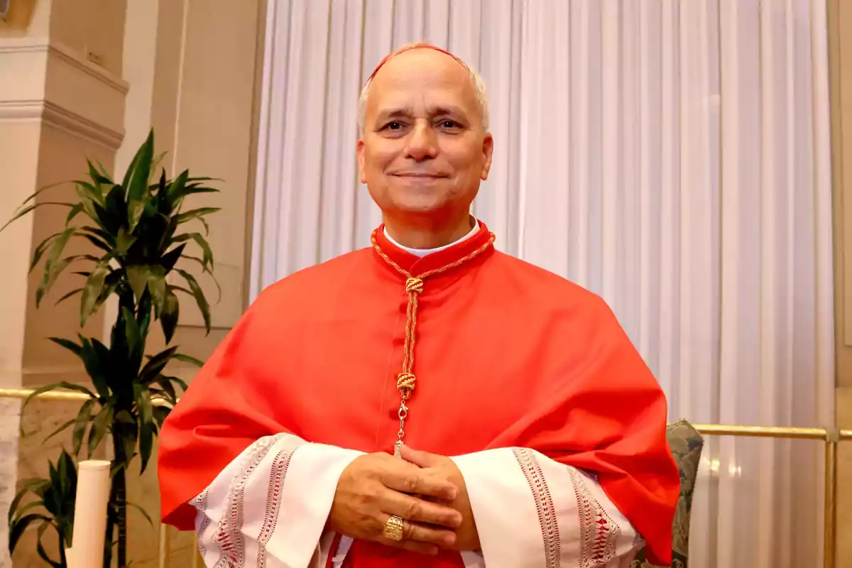 Un hombre con vestimenta religiosa roja y blanca posando frente a una cortina blanca con una planta al lado. Un hombre con vestimenta religiosa roja y blanca posando frente a una cortina blanca con una planta al lado.