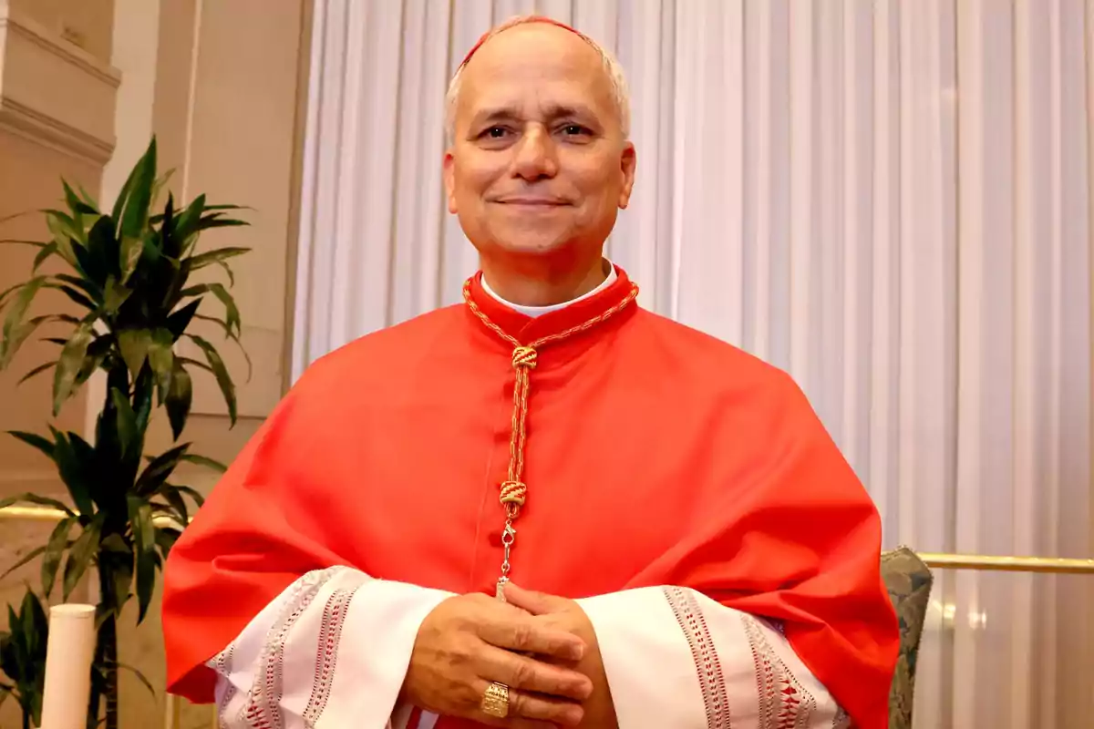 Un hombre con vestimenta religiosa roja y blanca sonríe frente a una cortina blanca con una planta al lado.