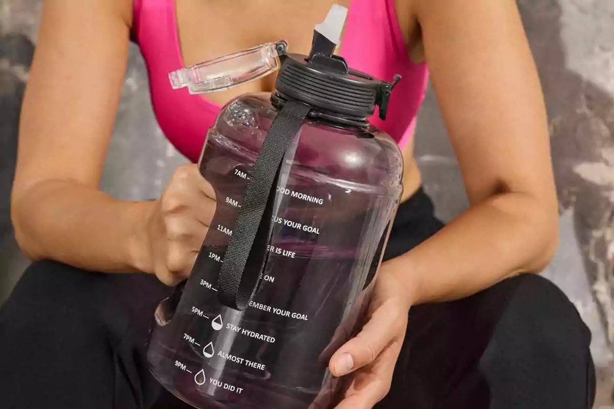 Una persona en ropa deportiva sostiene una botella grande de agua con marcas de tiempo y mensajes motivacionales. Una persona en ropa deportiva sostiene una botella grande de agua con marcas de tiempo y mensajes motivacionales.