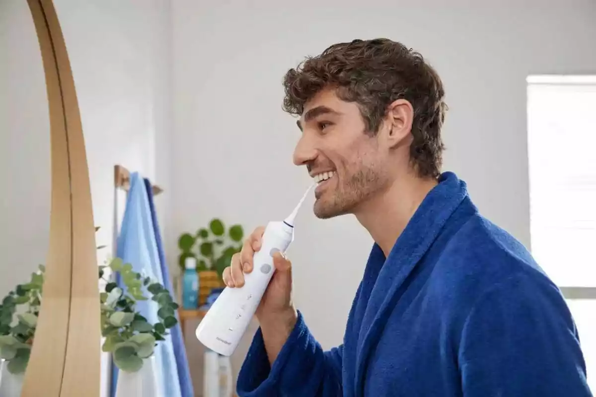 Hombre usando un irrigador dental frente al espejo en el baño, vistiendo una bata azul. Hombre usando un irrigador dental frente al espejo en el baño, vistiendo una bata azul.
