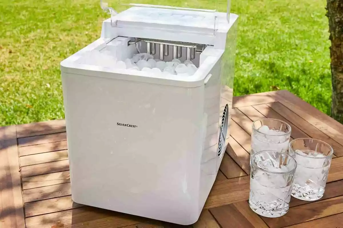 Máquina de hacer hielo blanca sobre una mesa de madera al aire libre con tres vasos llenos de hielo al lado. Máquina de hacer hielo blanca sobre una mesa de madera al aire libre con tres vasos llenos de hielo al lado.