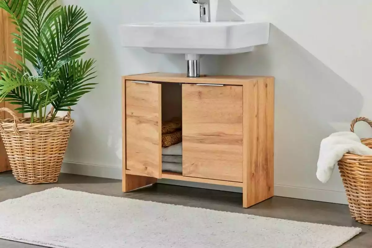 Un mueble de baño de madera clara con puertas abiertas mostrando toallas en su interior, ubicado debajo de un lavabo blanco, acompañado de una planta en una cesta de mimbre y una alfombra blanca en el suelo. Un mueble de baño de madera clara con puertas abiertas mostrando toallas en su interior, ubicado debajo de un lavabo blanco, acompañado de una planta en una cesta de mimbre y una alfombra blanca en el suelo.