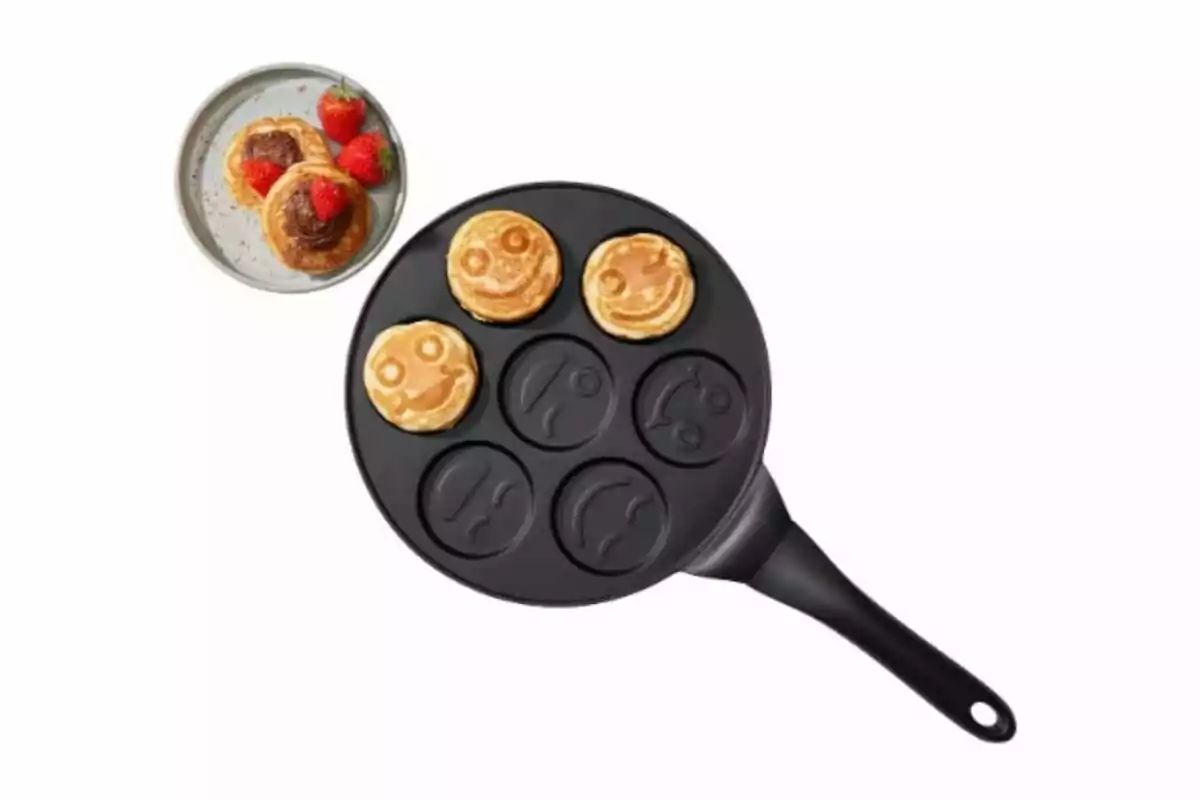 Sartén con moldes de caritas sonrientes y tres pancakes cocidos junto a un plato con pancakes y fresas.