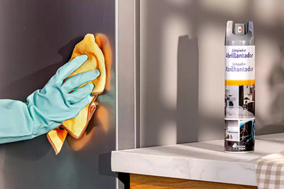 Persona limpiando una superficie de acero inoxidable con un paño amarillo y guantes, junto a un limpiador abrillantador en aerosol sobre una encimera. Persona limpiando una superficie de acero inoxidable con un paño amarillo y guantes, junto a un limpiador abrillantador en aerosol sobre una encimera.