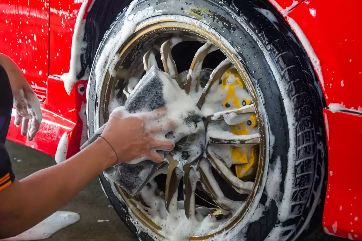 Persona lavando la llanta de un automóvil rojo con una esponja y jabón.