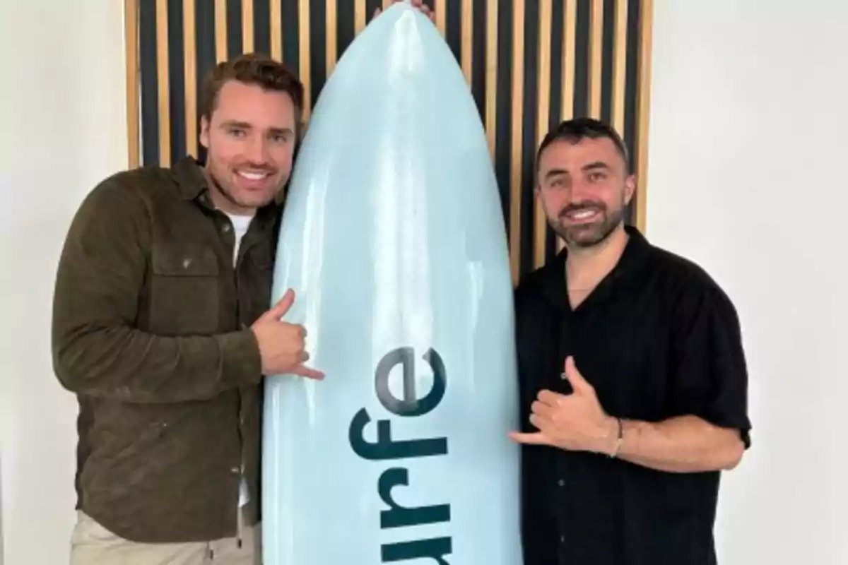 Dos hombres sonrientes posan junto a una tabla de surf azul claro con la palabra "surfe" escrita en ella.