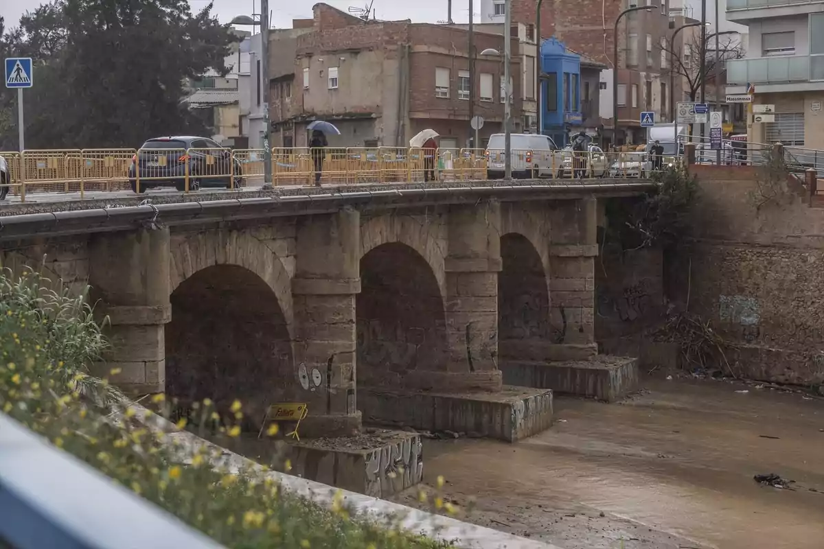 A stone bridge with arches over a dry riverbed on a rainy day, with people walking under umbrellas and cars driving by.