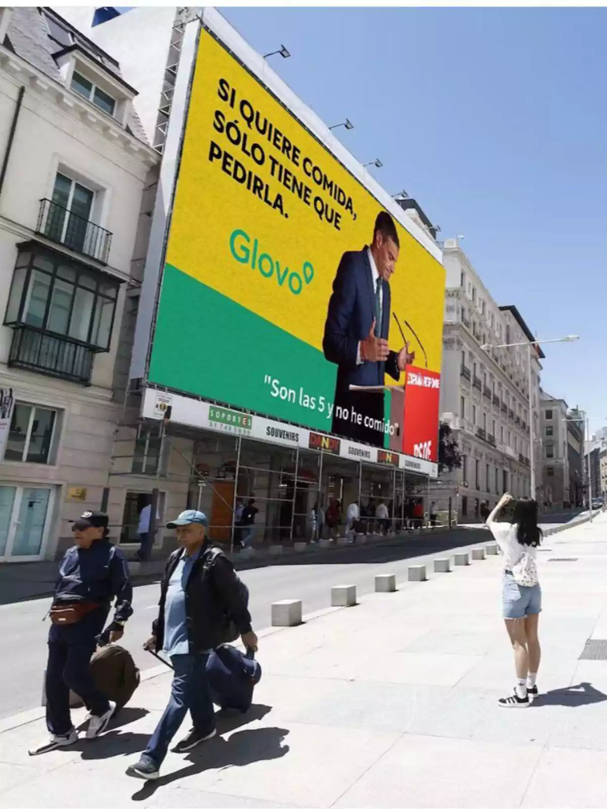 Un gran cartel publicitario de Glovo en una calle de ciudad muestra a un hombre de traje junto a un mensaje sobre pedir comida mientras varias personas caminan y una mujer toma una foto Un gran cartel publicitario de Glovo en una calle de ciudad muestra a un hombre de traje junto a un mensaje sobre pedir comida mientras varias personas caminan y una mujer toma una foto