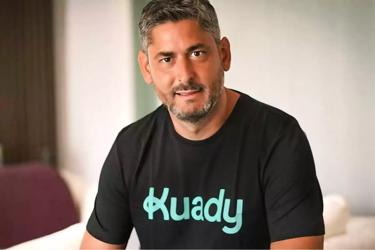 Hombre con barba y cabello corto canoso, vistiendo una camiseta negra con la palabra "Kuady" escrita en letras verdes.