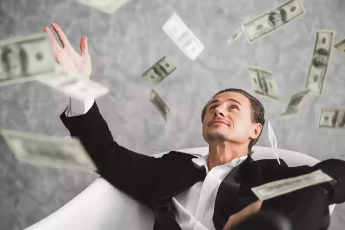 Hombre sentado en un sofá viendo hacia arriba con la mano alzada mientras caen billetes de dólar sobre él