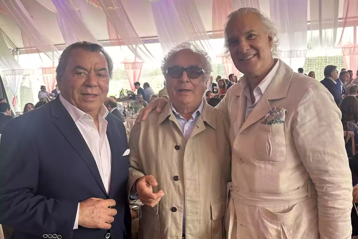 Tres hombres posando juntos en un evento social, con decoración de telas colgantes en el fondo.