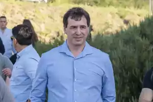 Hombre con camisa azul claro de pie al aire libre con personas y vegetación en el fondo.