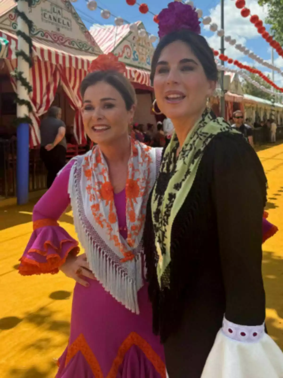 Lourdes Montes y otra mujer vestidas con trajes tradicionales posan sonrientes en un evento festivo al aire libre, con casetas decoradas y farolillos de colores en el fondo. Lourdes Montes y otra mujer vestidas con trajes tradicionales posan sonrientes en un evento festivo al aire libre, con casetas decoradas y farolillos de colores en el fondo.