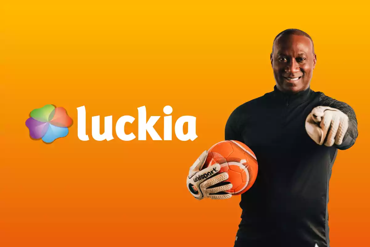 Hombre con guantes de portero sosteniendo un balón de fútbol frente a un fondo naranja con el logo de Luckia.