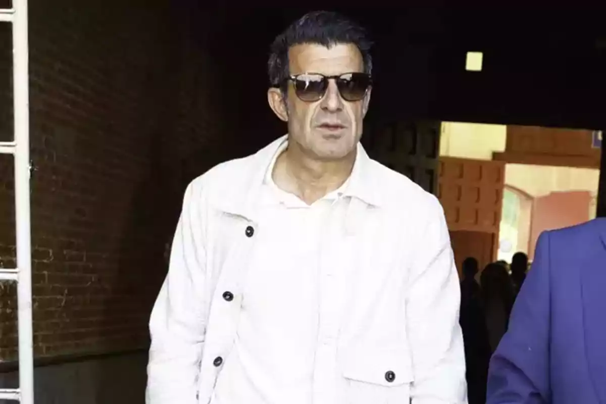 Luis Figo with sunglasses and a white jacket walks down a dark hallway. Luis Figo with sunglasses and a white jacket walks down a dark hallway.