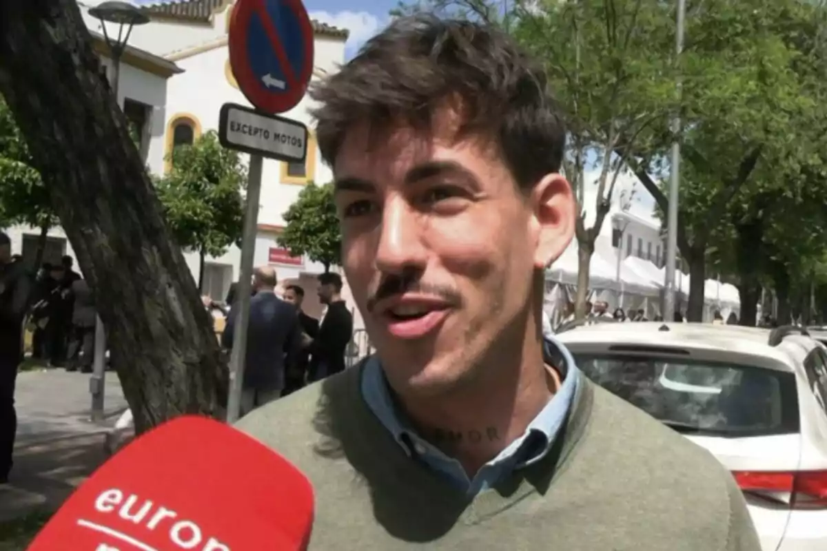 Luitingo sonriente es entrevistado en la calle, con un micrófono rojo en primer plano y un cartel de tráfico detrás. Luitingo sonriente es entrevistado en la calle, con un micrófono rojo en primer plano y un cartel de tráfico detrás.