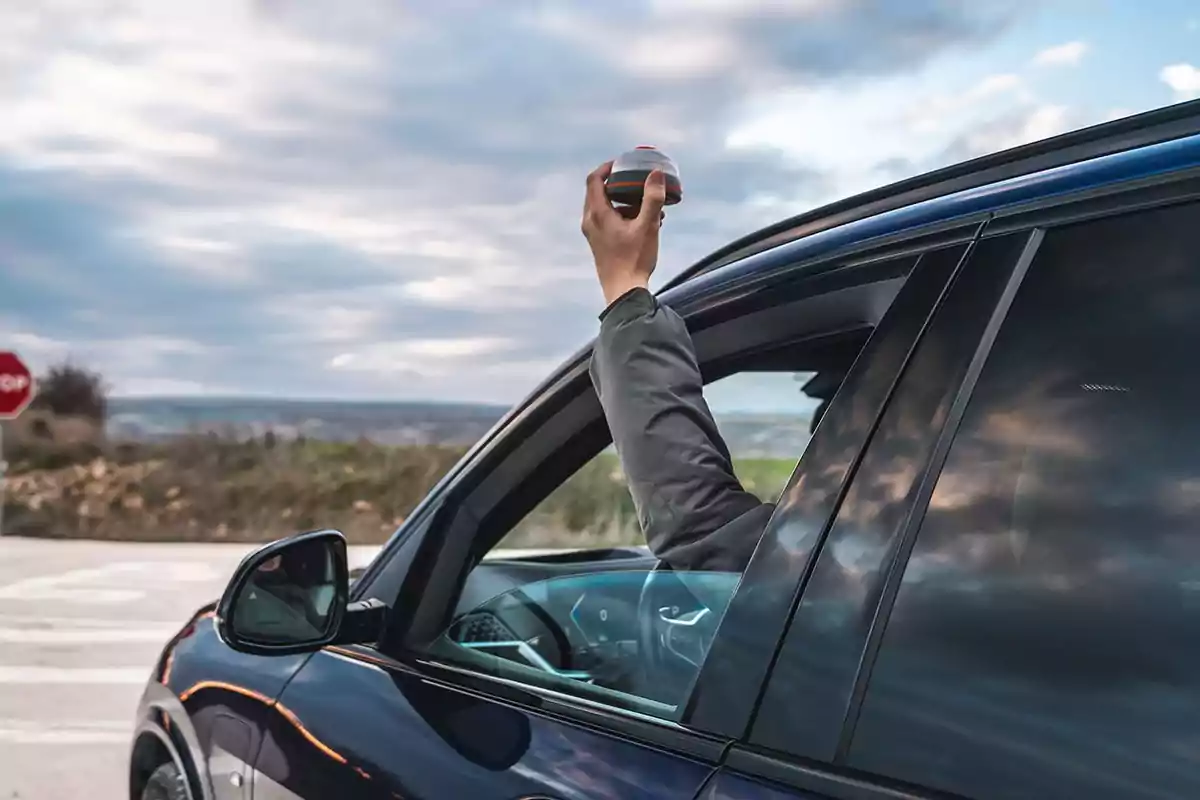 Persona saca la mano por la ventana de un auto sosteniendo un objeto redondo en un entorno al aire libre con cielo nublado.