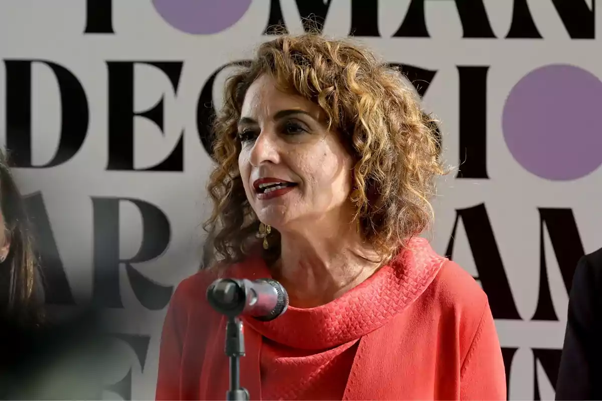 Una mujer de cabello rizado y castaño claro, vestida con una blusa roja, habla frente a un micrófono con un fondo de letras grandes y círculos morados.