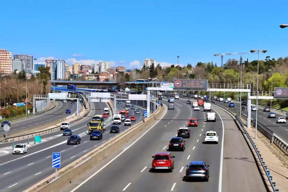 Autopista urbana con varios carriles y tráfico moderado, rodeada de edificios y áreas verdes bajo un cielo despejado.