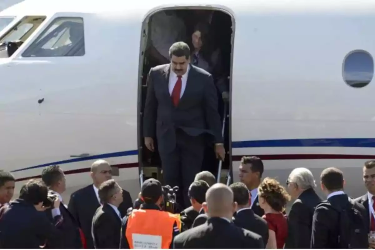 Un hombre con traje y corbata roja baja de un avión rodeado de varias personas.