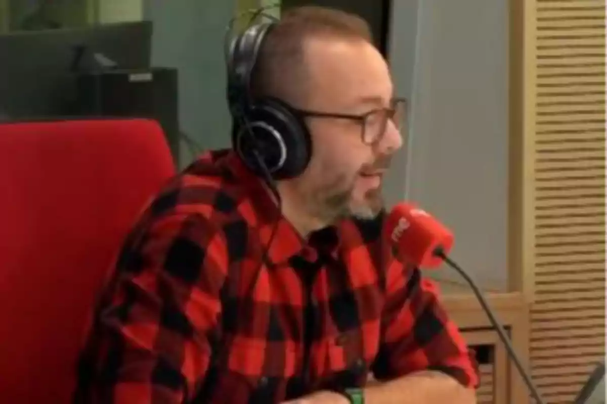 Un hombre con camisa de cuadros rojos y negros está hablando en un micrófono de radio, usando auriculares y sentado en una silla roja. Un hombre con camisa de cuadros rojos y negros está hablando en un micrófono de radio, usando auriculares y sentado en una silla roja.