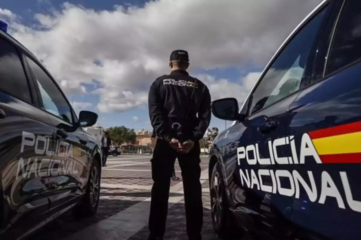 Un agente de la Policía Nacional está de pie entre dos coches patrulla en una plaza, con las manos detrás de la espalda y el cielo parcialmente nublado.