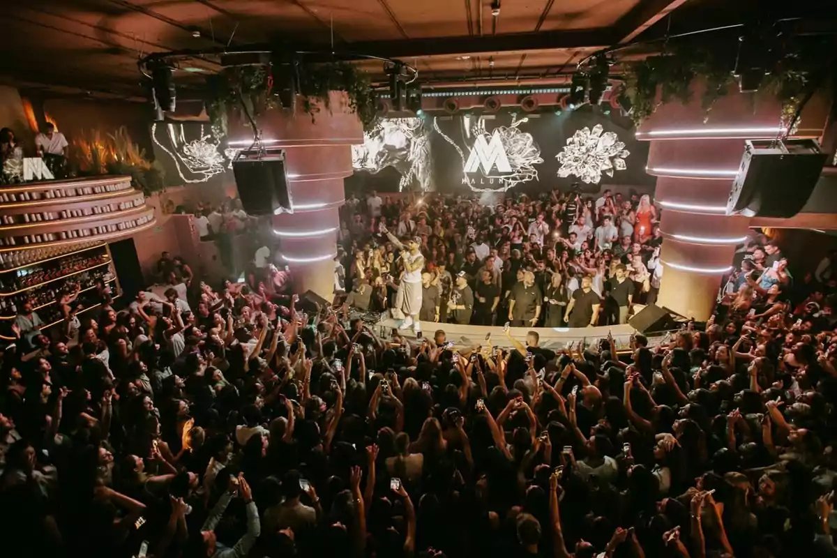 Una multitud de personas en un concierto en un club nocturno con un escenario iluminado y un artista actuando mientras el público levanta sus teléfonos móviles para grabar.