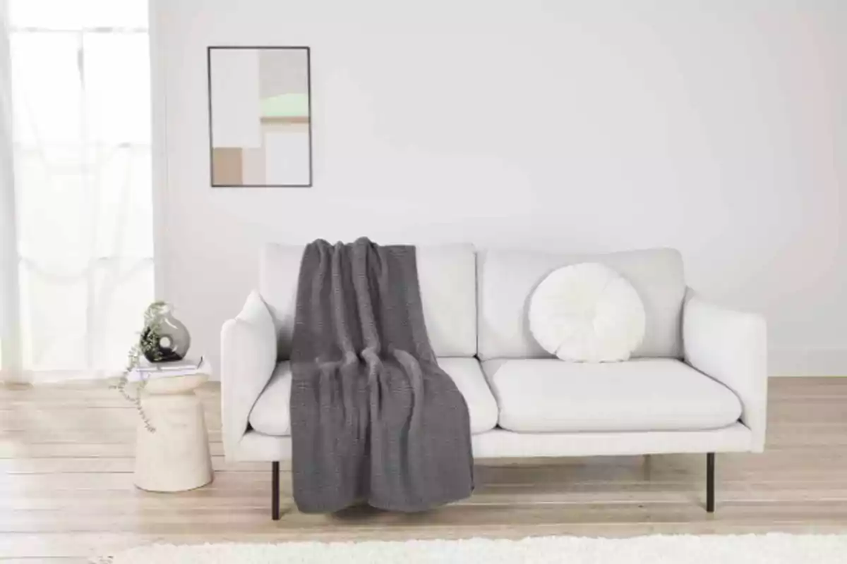 Un sofá blanco con una manta gris y un cojín redondo, junto a una mesa auxiliar con libros y un jarrón, en una sala de estar minimalista con un cuadro en la pared. Un sofá blanco con una manta gris y un cojín redondo, junto a una mesa auxiliar con libros y un jarrón, en una sala de estar minimalista con un cuadro en la pared.